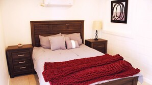 Un lit bien fait, avec une couverture rouge, une tête de lit en bois et une table de chevet surmontée d’une lampe.
