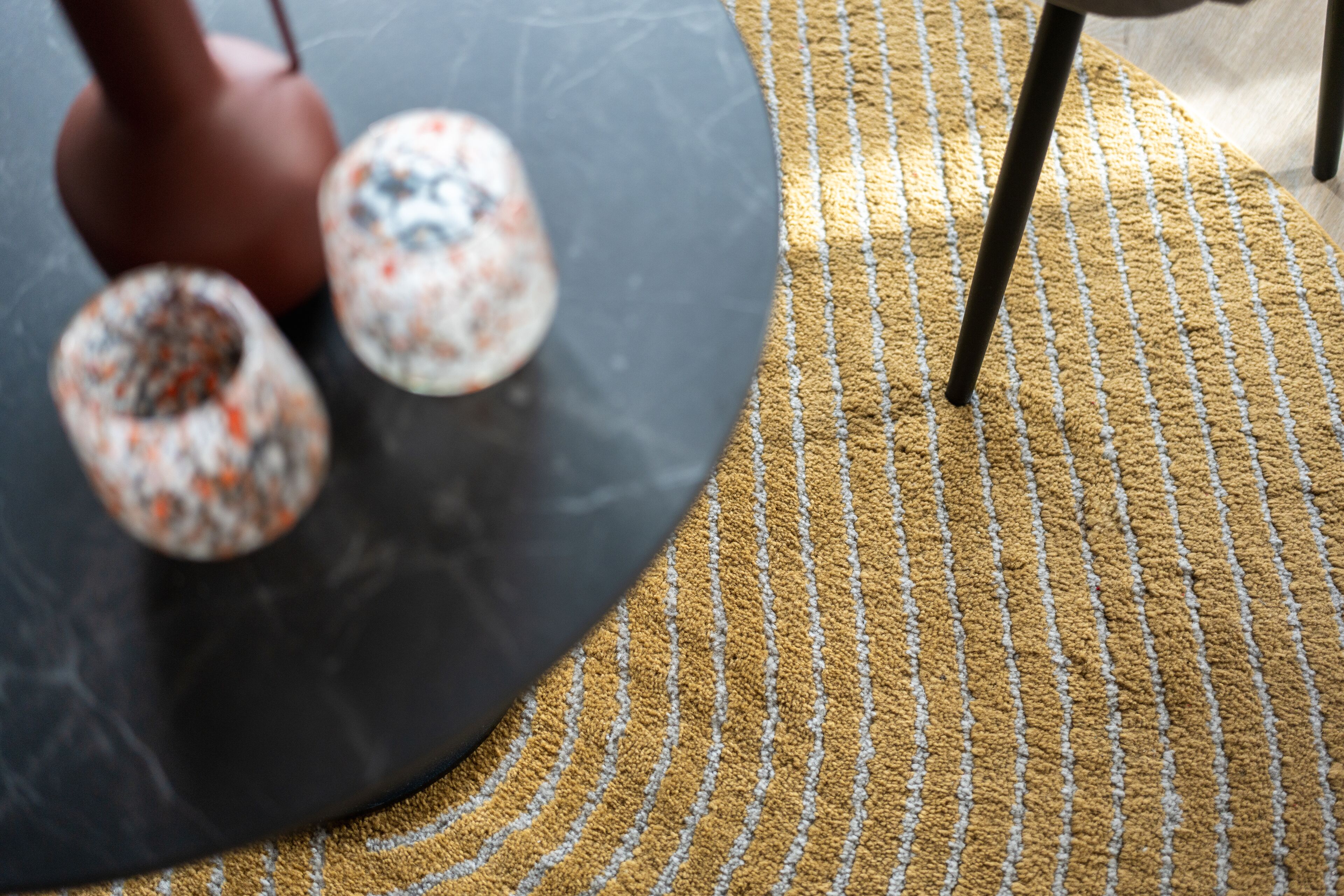 Un tapis à motifs texturés, une table sombre aux bords incurvés et un vase marron orné de motifs floraux.