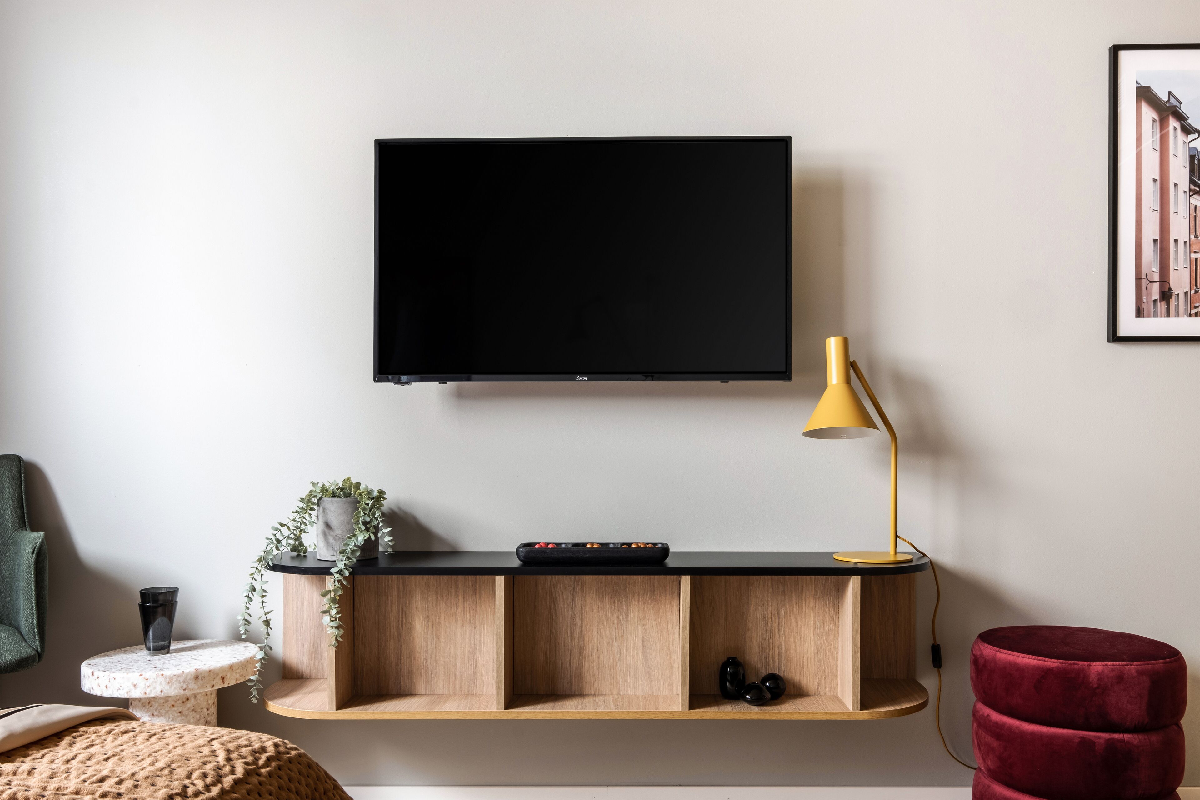 Una sala de estar moderna con un televisor montado, una mesa consola de madera, un reposapiés rojo y una lámpara de pie amarilla.
