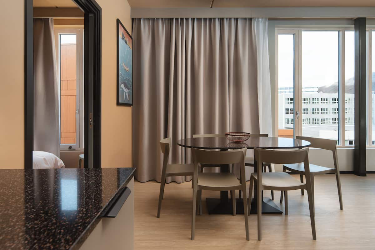A hotel room with a dining area featuring a round table and four chairs, a large window with curtains, and a view of buildings outside.