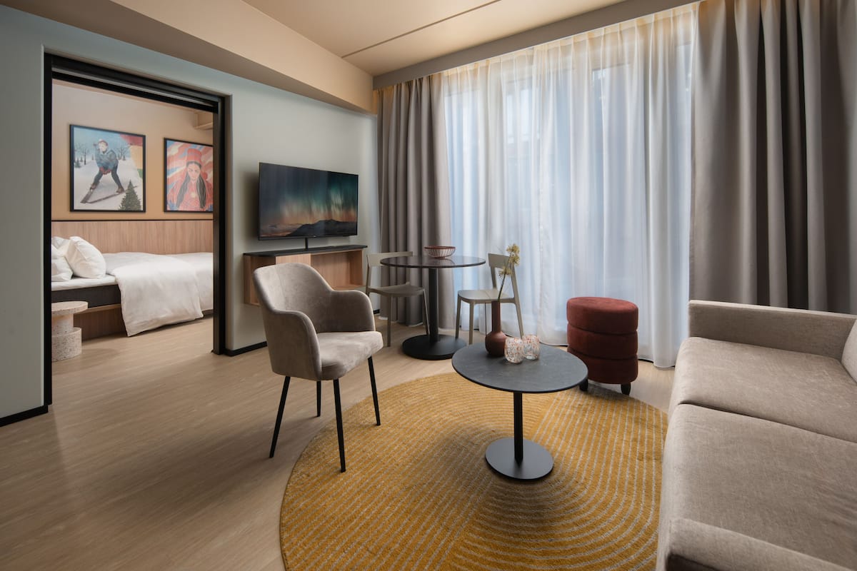 A modern hotel room with a bed, two armchairs, a small round table, a sofa, a TV, and a large window with curtains.