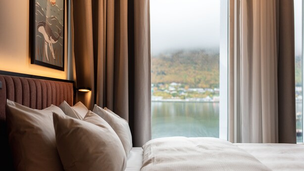 A hotel room with a bed, pillows, a headboard, a painting on the wall, and a view of a body of water and hills. - Skaret by Vander (Tromsø)
