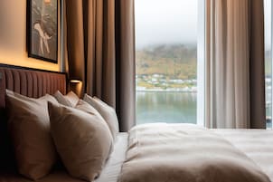 Habitación de hotel con cama, almohadas, cabecera, un cuadro en la pared y vista a un cuerpo de agua y colinas.