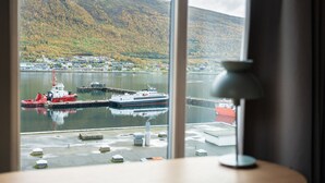 A view of a harbor with a tugboat, a large ship, and a town across the water, framed by a window with a lamp on a table. - Skaret by Vander (Tromsø)