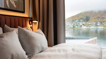 A hotel room with a bed, pillows, a headboard, a wall-mounted lamp, and a window with a view of a lake and buildings.