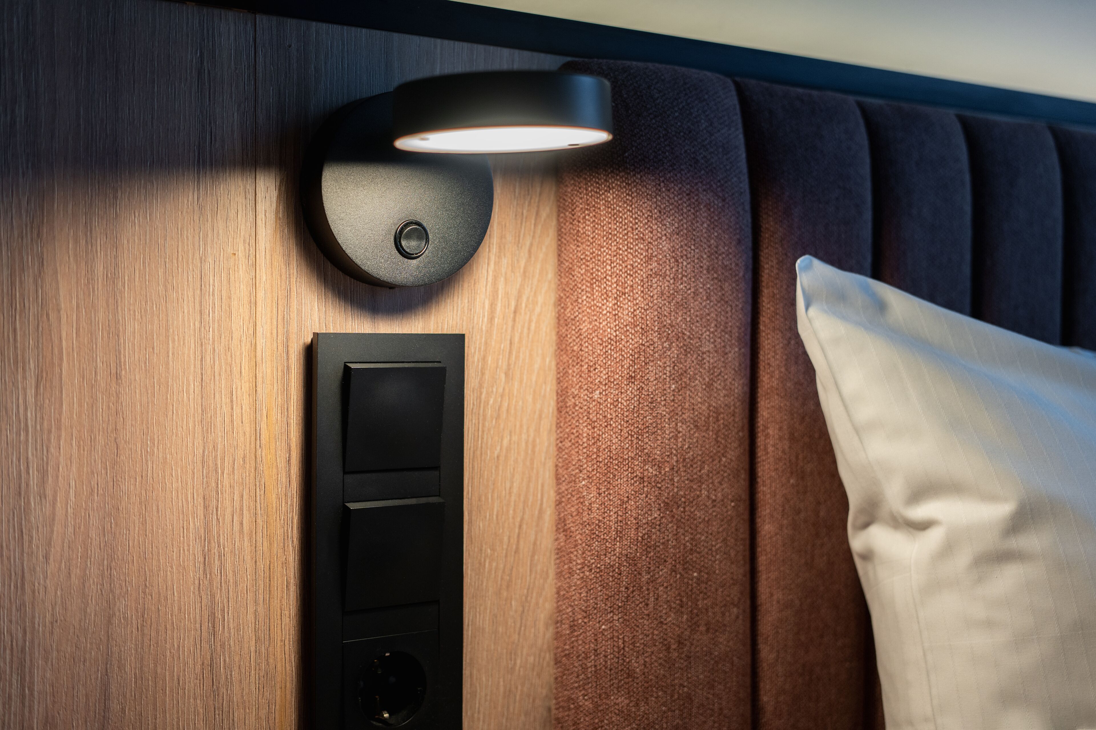 A wall-mounted light fixture above a control panel, a white pillow, and a textured wall.