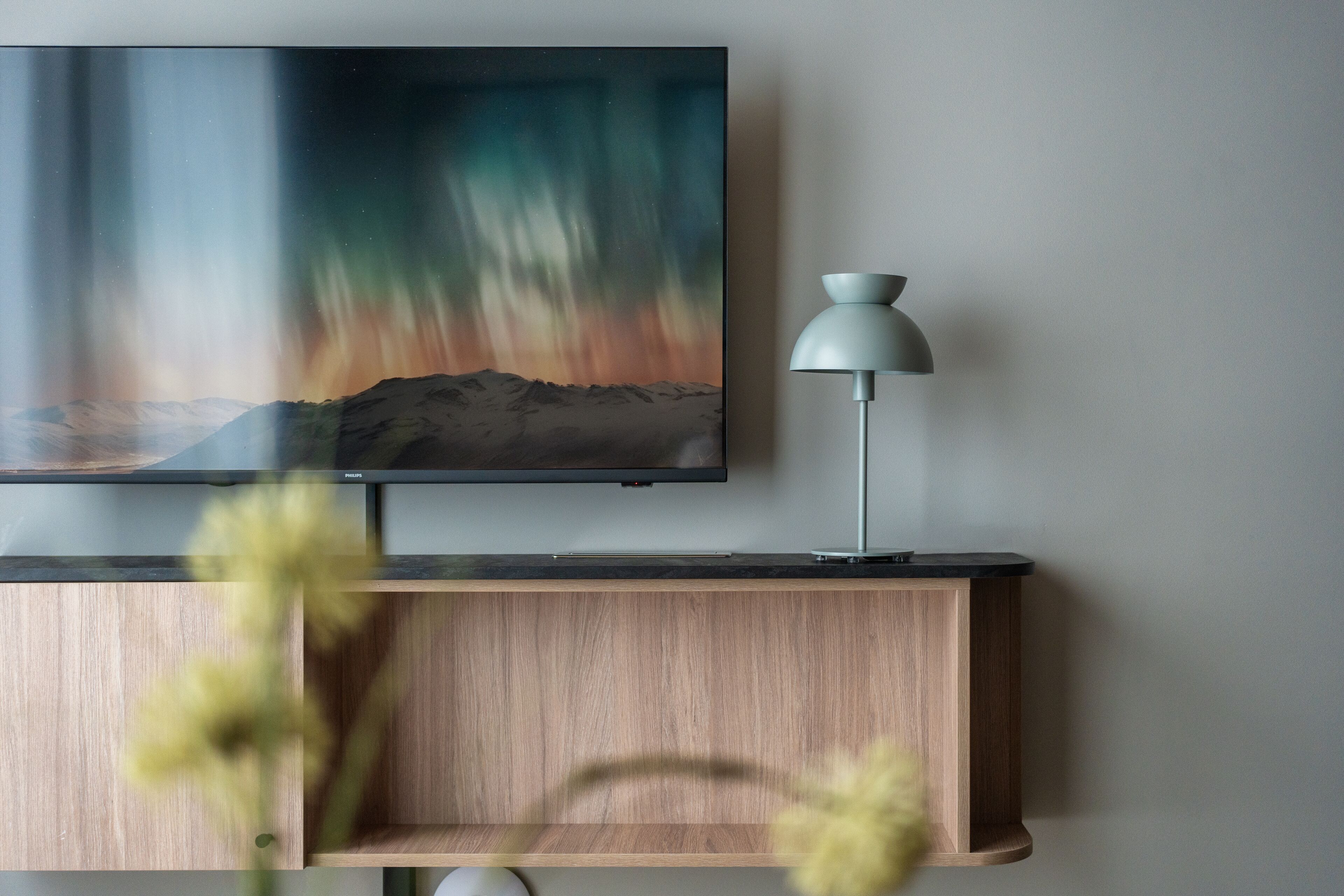 A flat-screen TV mounted on a wooden shelf, with a modern lamp beside it.