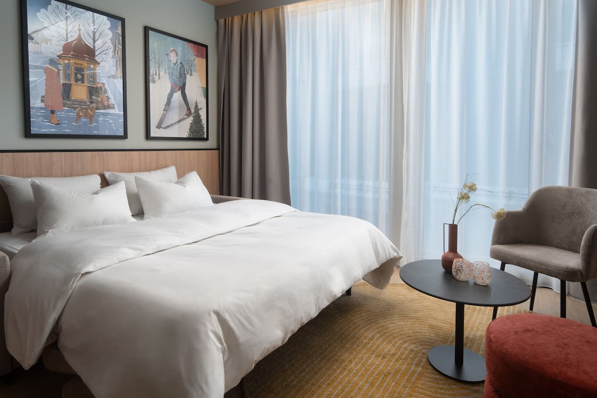 A hotel room with a large bed, a small round table with a vase, a chair, and two framed pictures on the wall.