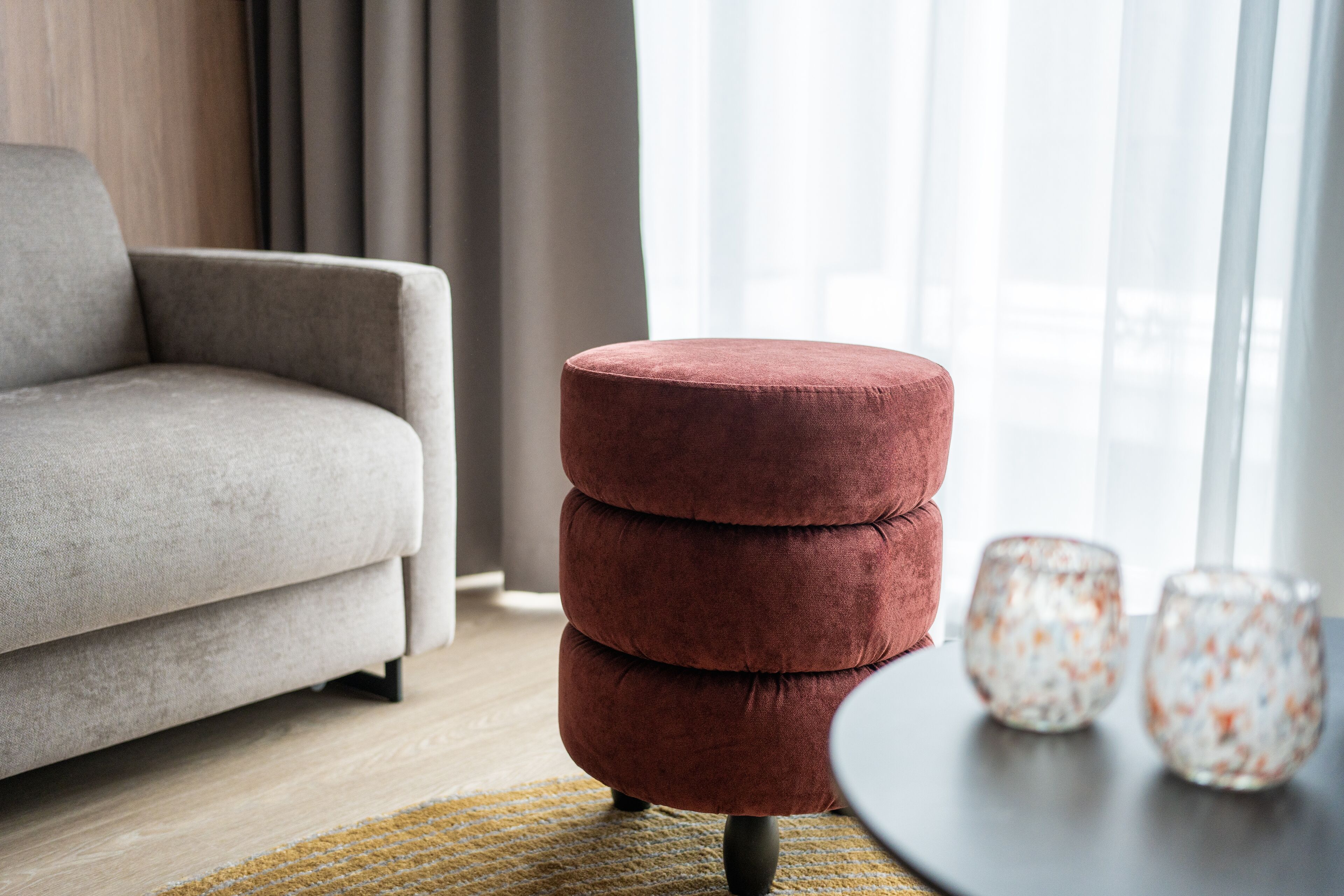 A modern living room with a grey sofa, a stack of red ottomans, and two glass vases on a wooden floor.