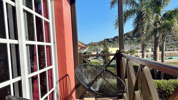 A balcony with two black metal chairs and a small table, overlooking a landscaped area with palm trees and a distant hill. - Spacious Villa in Quiet Location Close to Resorts (Buzanada)