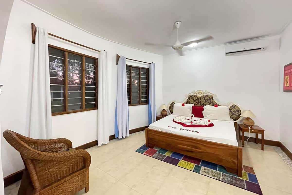Un dormitorio con cama, ventilador de techo, ventana con cortinas, una silla de mimbre y una alfombra colorida.