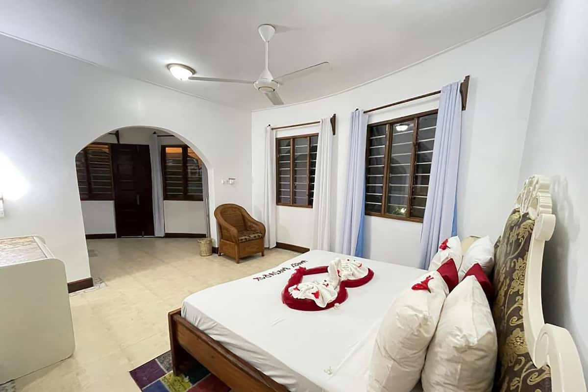 Un dormitorio con cama, una silla, ventilador de techo y ventana con cortinas.