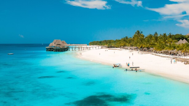 A beach with clear turquoise water, a white sandy shore, and a thatched-roof structure extending into the water. - Five Palms Bwejuu (Bwejuu)