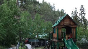 A playground with a wooden playhouse, a green slide, and various play equipment. - Timber Haven Cabins (Hill City)