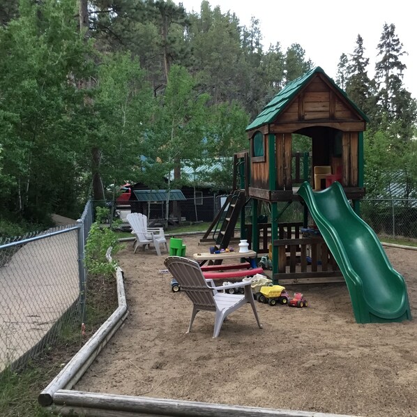 A playground with a wooden playhouse, a green slide, and various play equipment. - Timber Haven Cabins (Hill City)