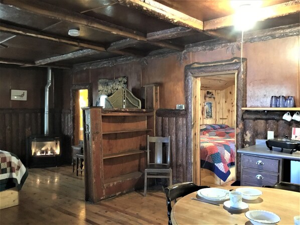 A cozy room with a wooden interior, a bed with a colorful quilt, a small table with plates and cups, and a chair. - Timber Haven Cabins (Hill City)