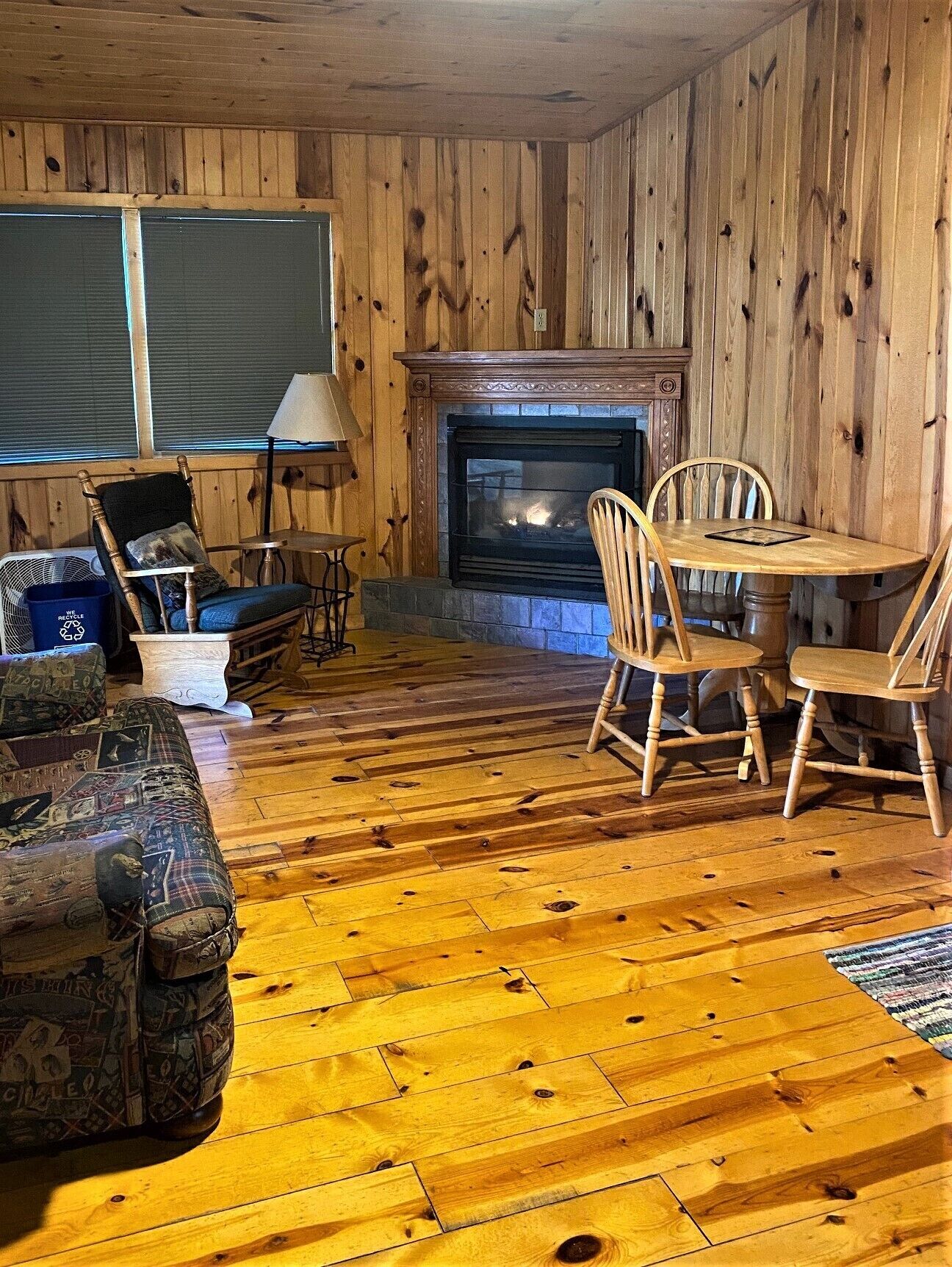 A cozy cabin interior with a wooden floor, a fireplace, a dining table with chairs, and a patterned couch.