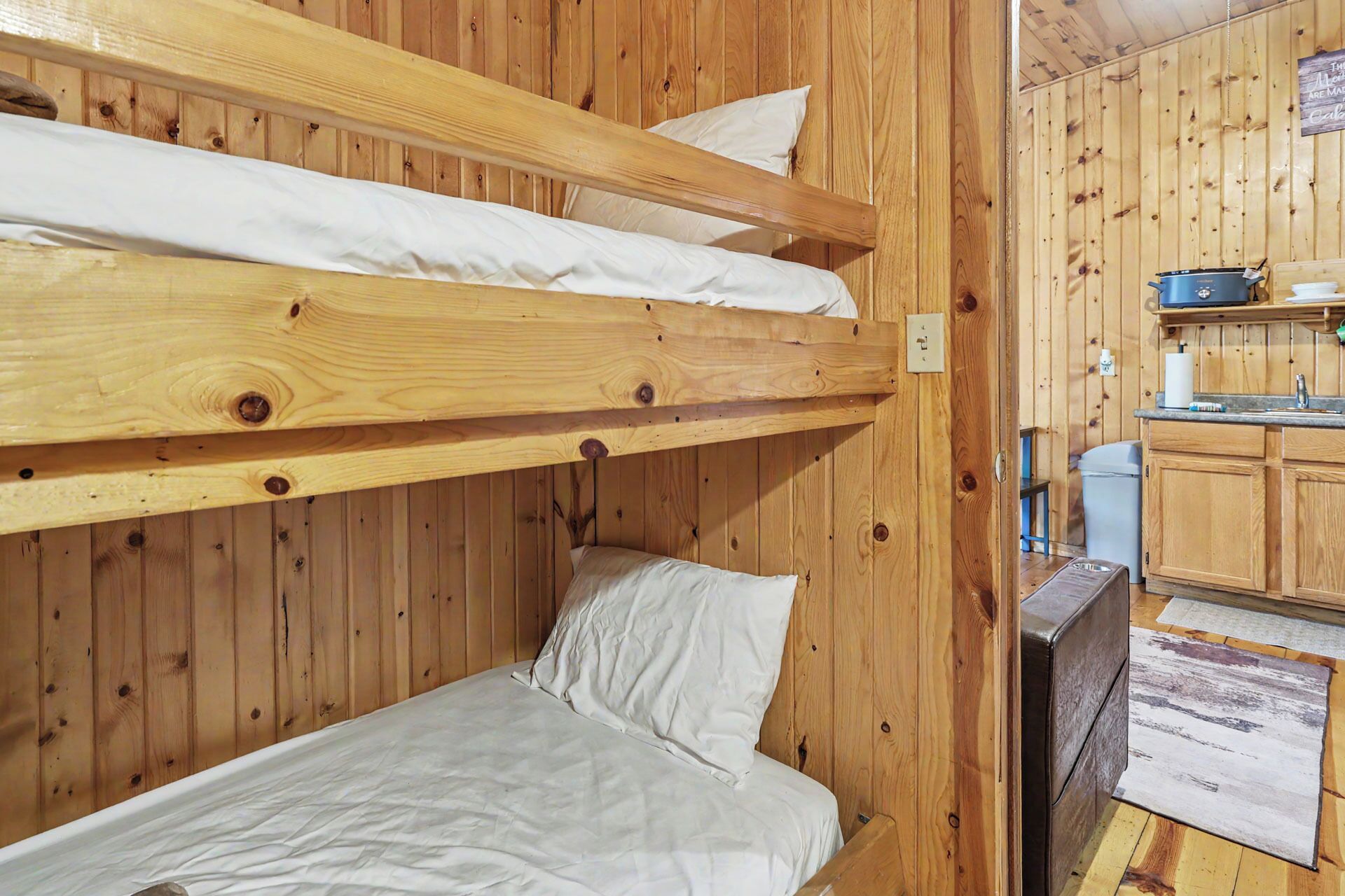 A bunk bed with white bedding, a wooden wall, and a kitchenette with a sink and stove.