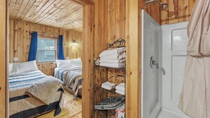 A wooden cabin with two beds, a metal rack with towels, a shower, and a window with curtains. - Timber Haven Cabins (Hill City)
