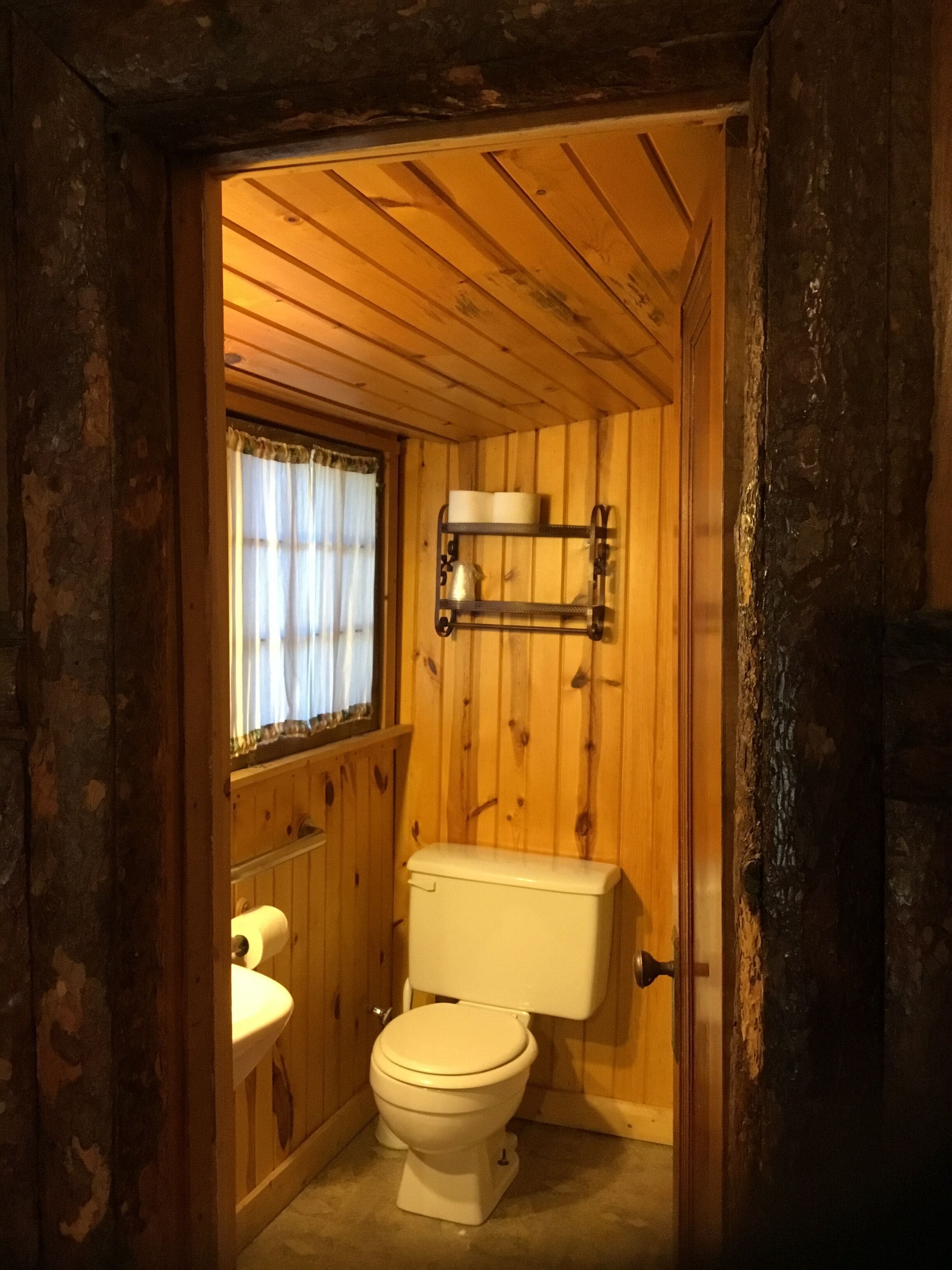 A small, wooden-paneled bathroom with a toilet, a sink, and a window with curtains.
