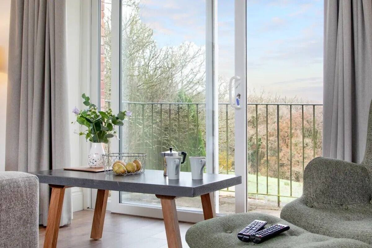 A living room with a sofa, a green armchair, a table with a vase of flowers, and a balcony with a view of trees and a fence.