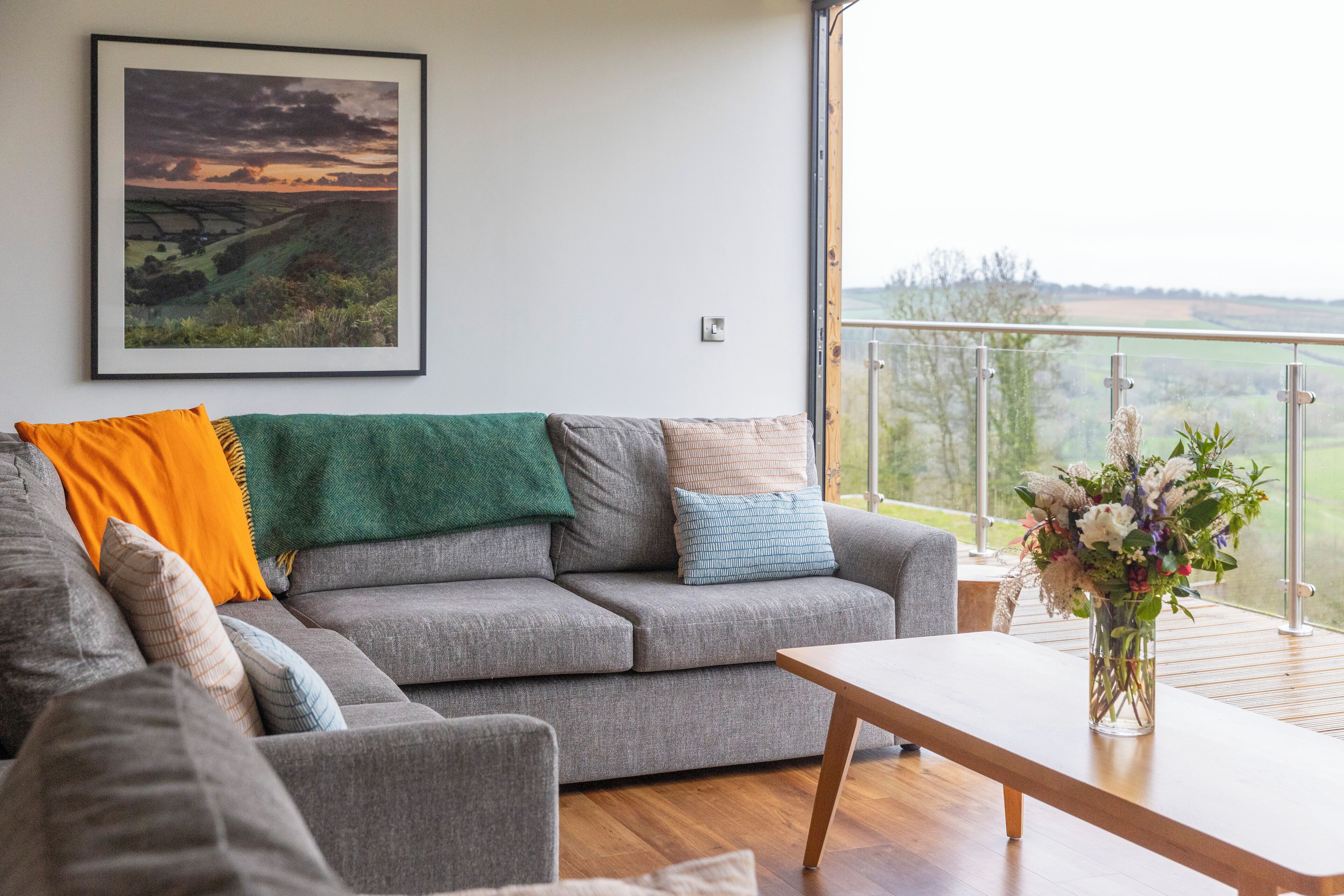 Een moderne woonkamer met een grijze bank, een houten salontafel en een lijstwerk met een landschapsdecoratie aan de muur.