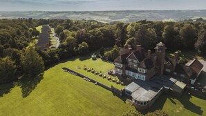 Une vue aérienne d’un vaste domaine comprenant une imposante maison de style Tudor, de vastes pelouses verdoyantes et un ensemble de bâtiments plus petits.