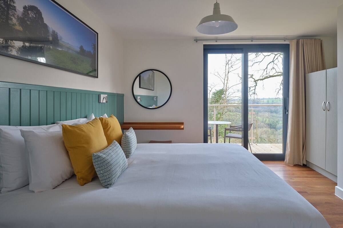 A bedroom with a large bed, a view of the outdoors through a sliding glass door, a round mirror, and a painting on the wall.