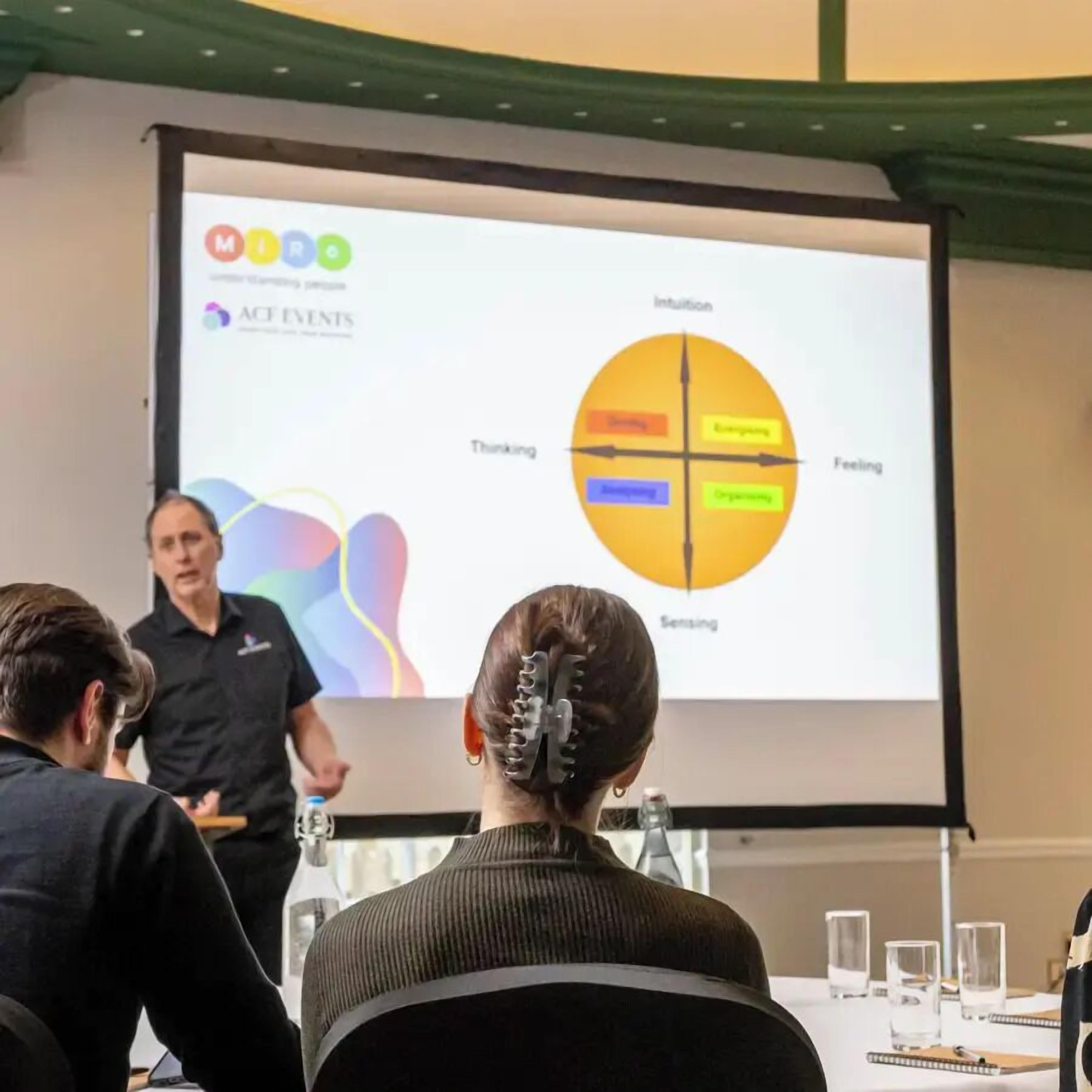 Une présentation avec un diagramme circulaire affiché sur un écran, une personne qui fait la présentation et un public assis.