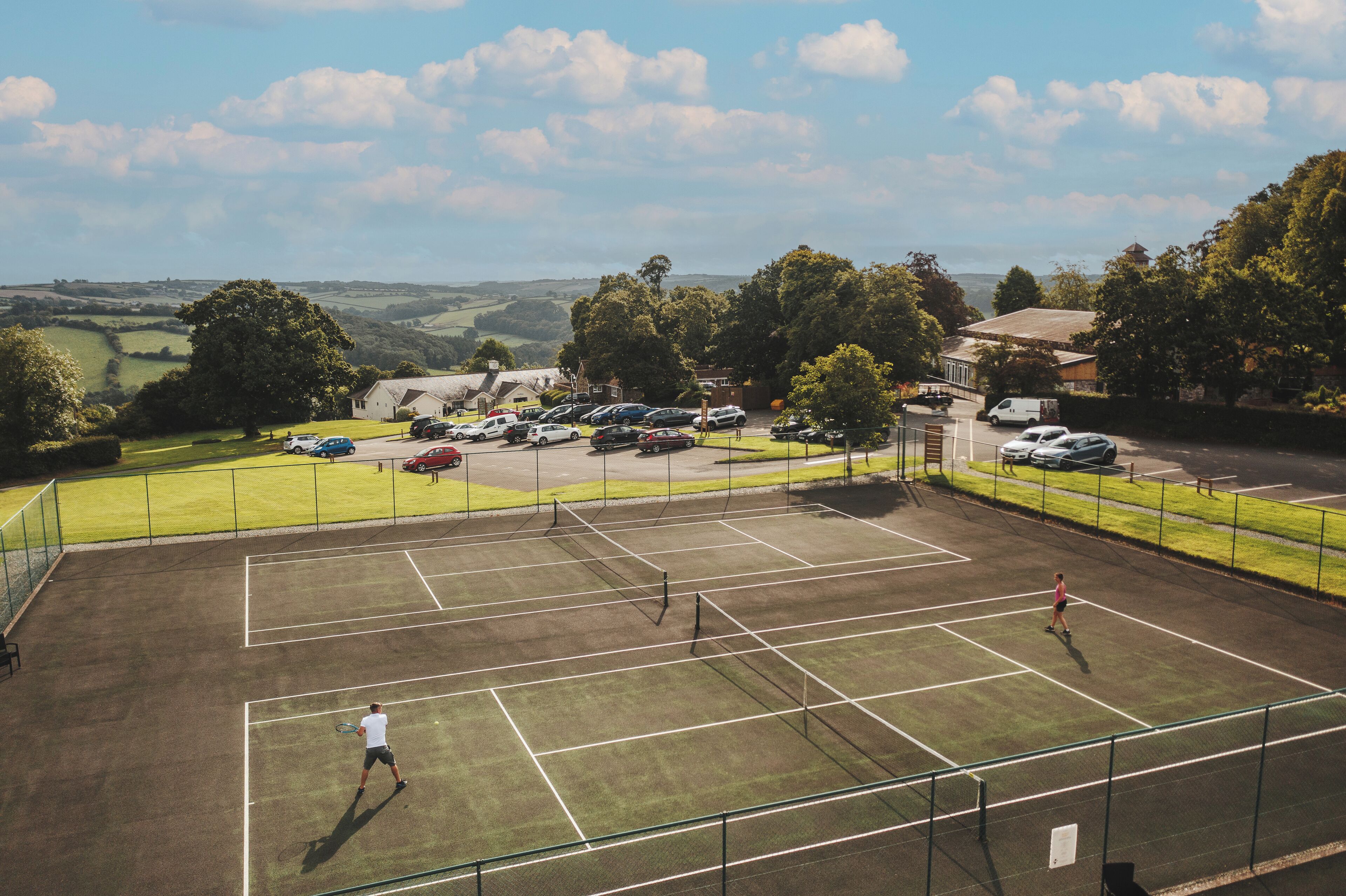 A tennis court with a net, surrounded by a parking area and buildings.