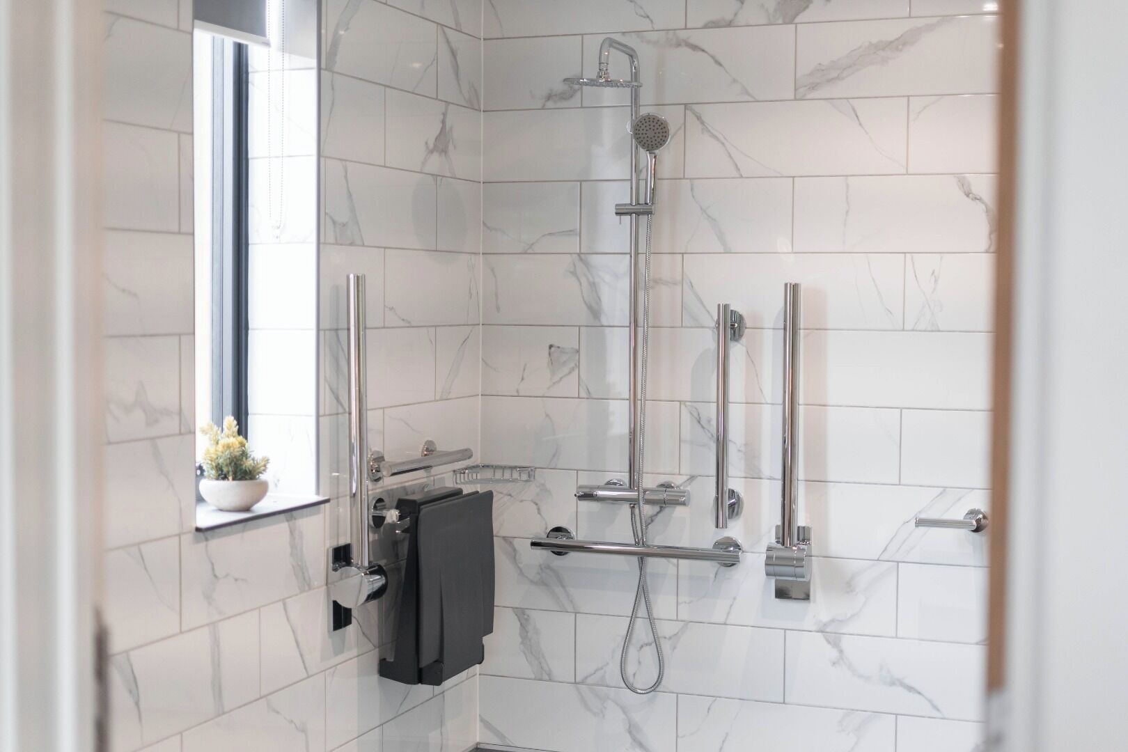 Een moderne badkamer met witte marmertegeltjes, een douchecabine van glas, een handdouche, een handdoekrek en een potplant.