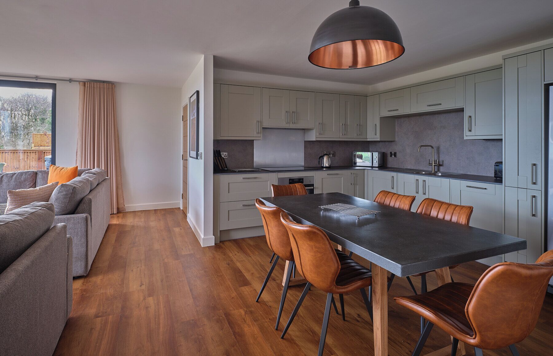 A modern kitchen with grey cabinets, a dark countertop, and a large table with leather chairs.