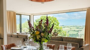 Un coin repas avec une grande table dressée pour six personnes, un luminaire suspendu moderne et une vue sur la campagne.