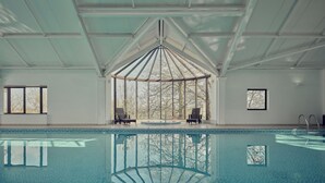 Une piscine intérieure avec des parois en verre transparent, un plafond haut et un espace central aménagé avec des fauteuils, offrant une vue dégagée.