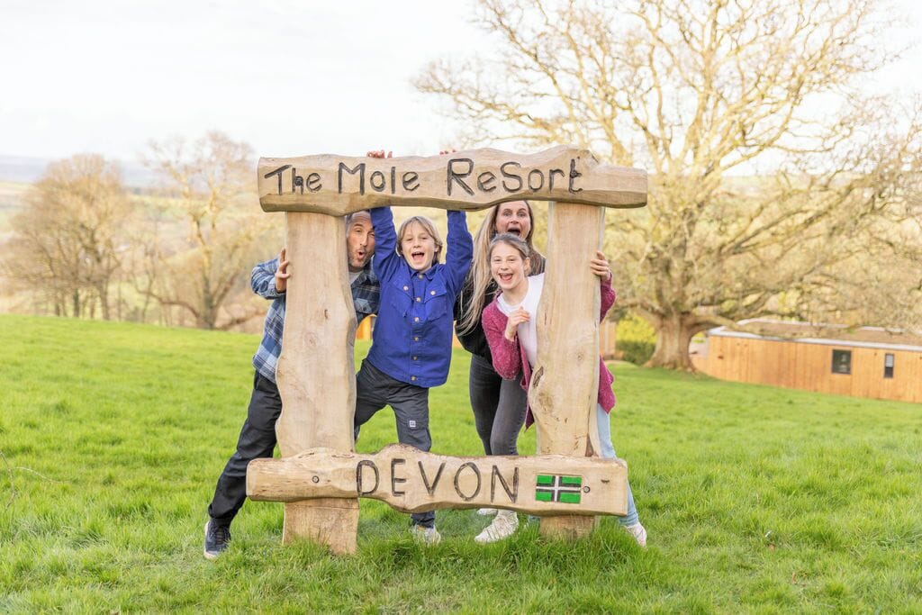 A wooden sign with "The Mole Resort" and "DEVON" written on it, held by people in a grassy field.