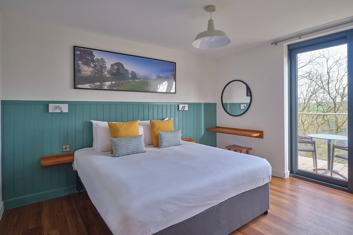 A bedroom with a large bed, a painting, a round mirror, and a view of the outdoors through a large window.
