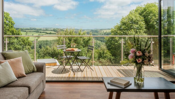 Un balcon spacieux doté d’un plancher en bois, d’un canapé, d’une table à manger et offrant une vue sur un paysage verdoyant.