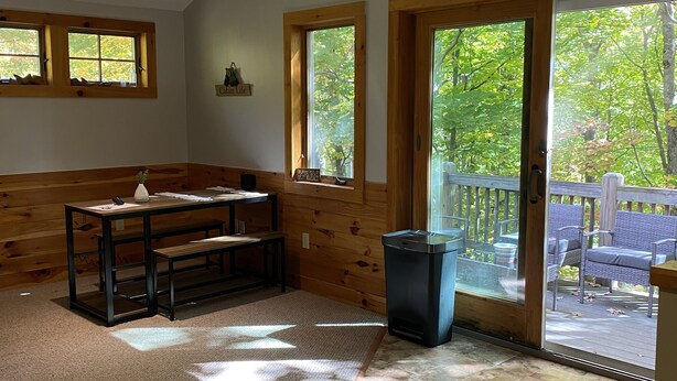 A room with a wooden bench, a table, and a trash can. There are windows and a door leading to an outdoor area with a wooden deck. - Lake & Mount Sunapee Apartment Getaway (Newbury)