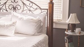 A neatly made bed with a white quilt, a bedside table with a lamp, and a window with blinds. - That Corner Lot-Charming Downtown Home w/Side Yard (Troy)