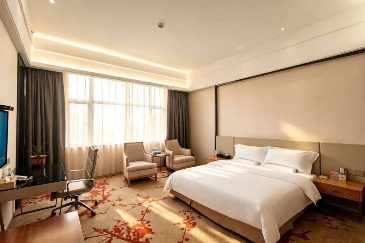 Habitación de hotel con cama, escritorio, sillas, televisor y un ventanal grande.