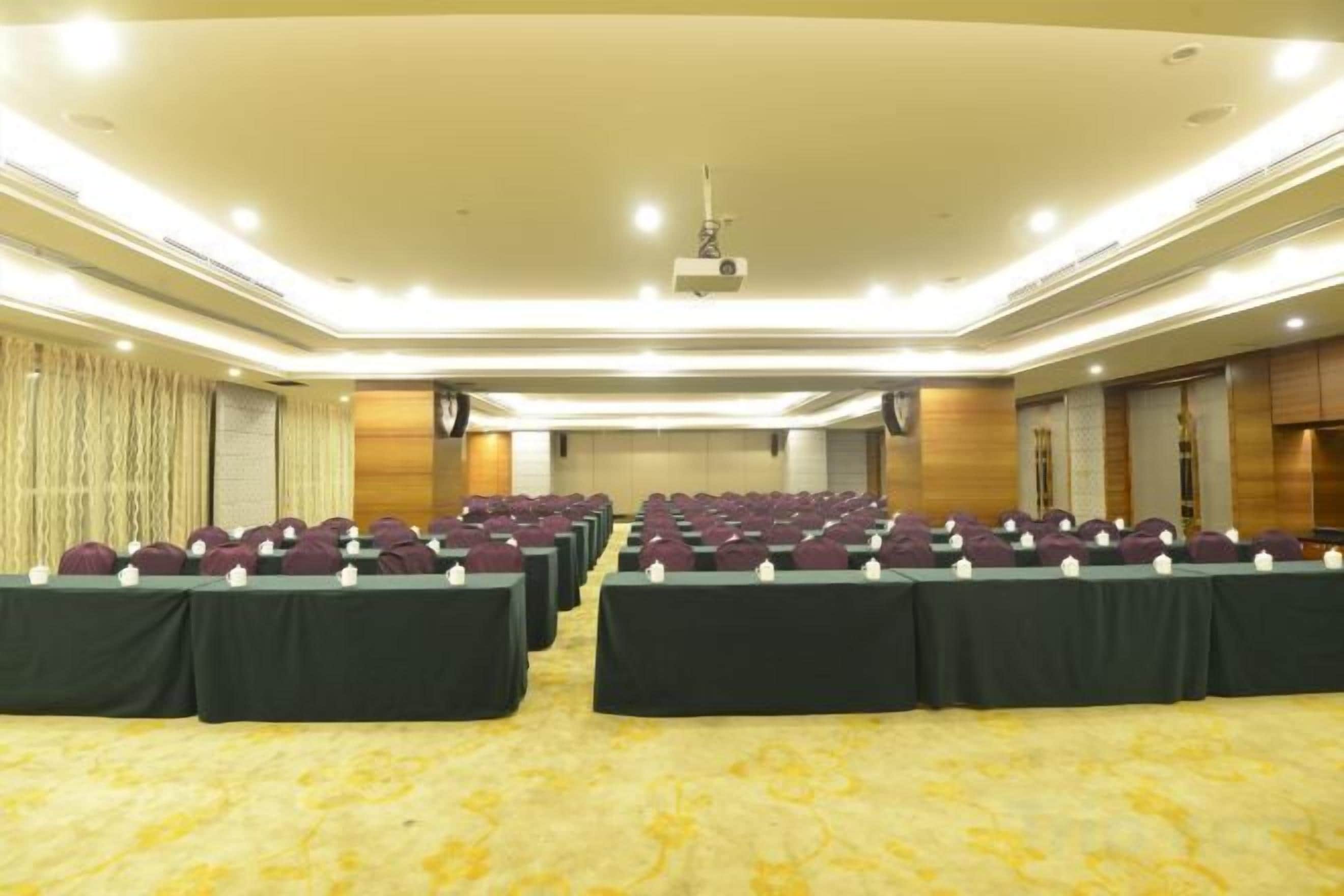 Un salón de conferencias con filas de mesas y sillas, un proyector y suelo alfombrado.