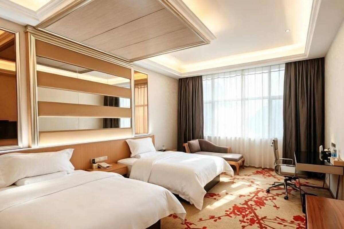 Habitación de hotel con dos camas, un escritorio, una silla, un ventanal con cortinas y un espejo sobre la cama.