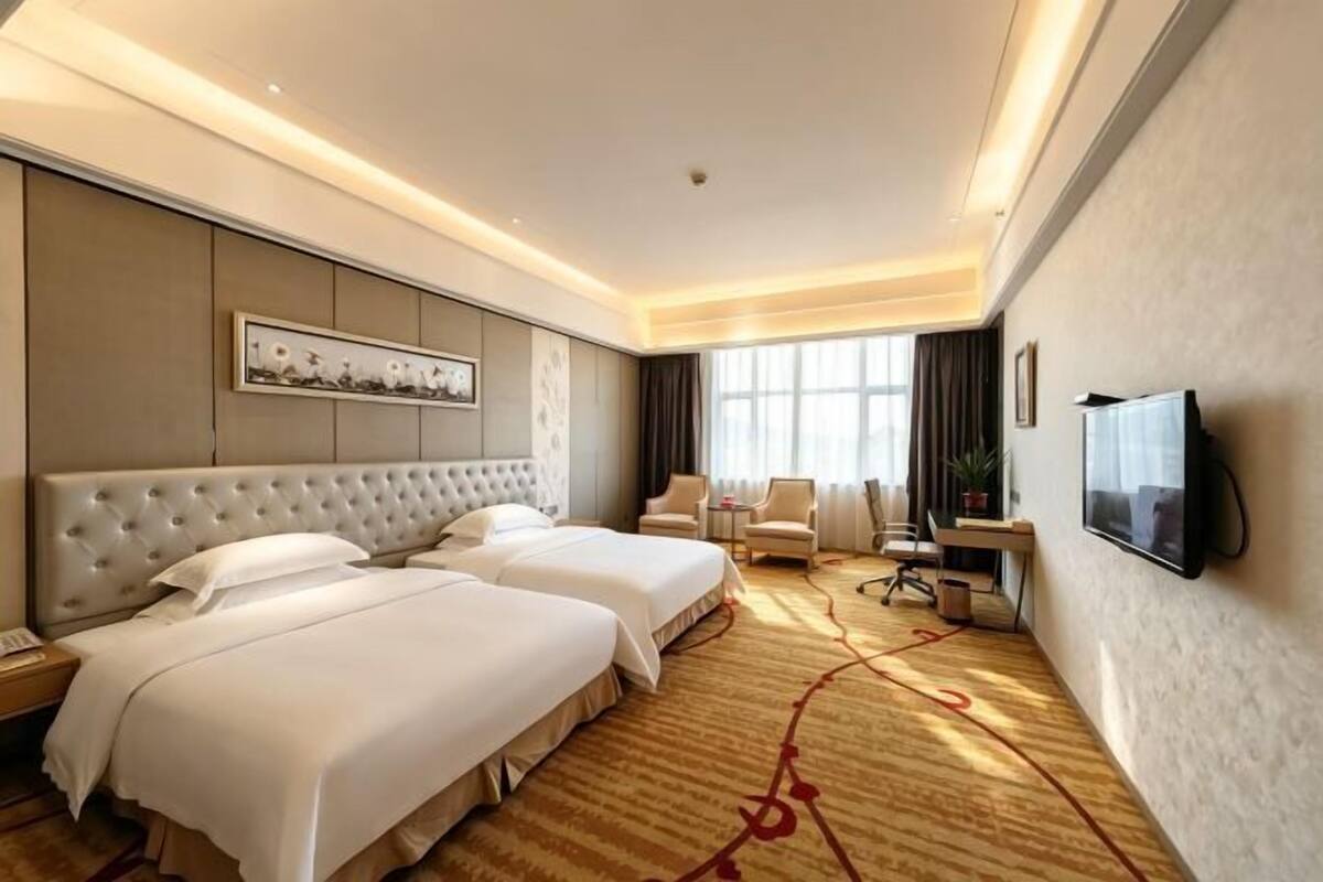 Habitación de hotel con dos camas, televisión, escritorio y ventana grande.