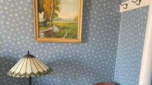 Habitación de esquina con papel tapiz azul estampado, una silla de madera con cojín verde, una mesita con lámpara y un cuadro enmarcado en la pared.
