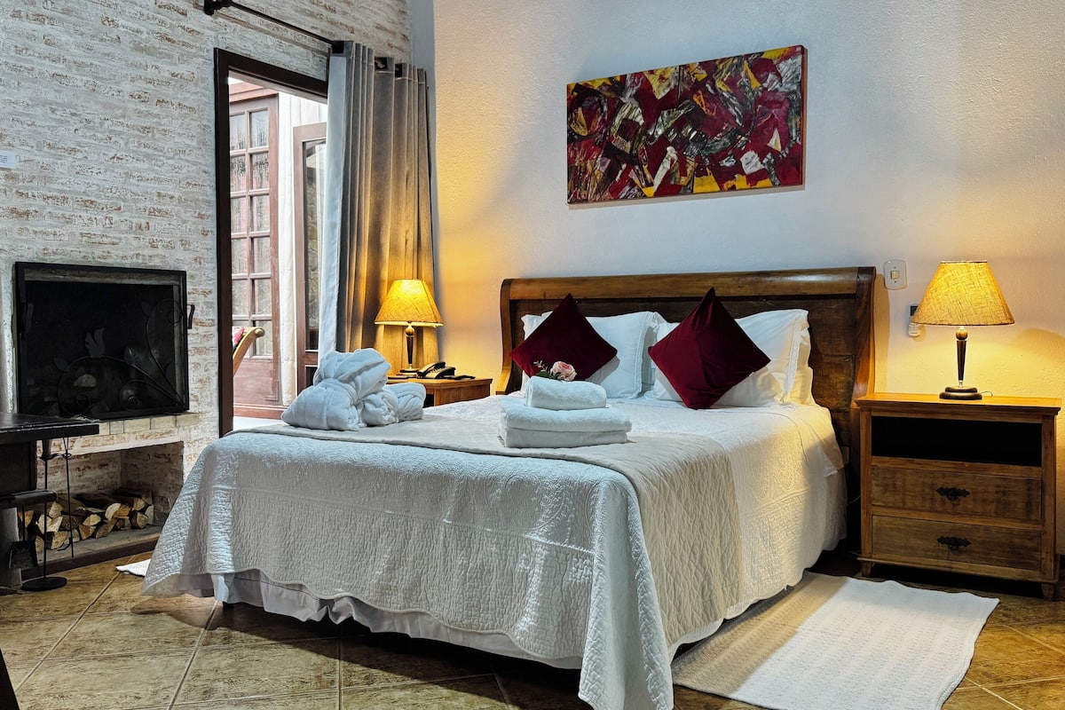 Quarto de hotel com uma cama grande, duas mesas de cabeceira com luminárias, uma TV, uma lareira e um quadro abstrato colorido na parede.