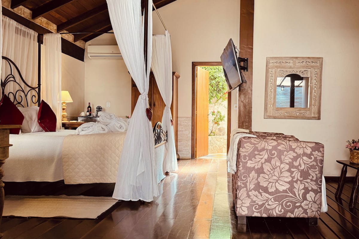Quarto com cama, mesa de cabeceira com abajur, televisão na parede e janela com vista para o verde.