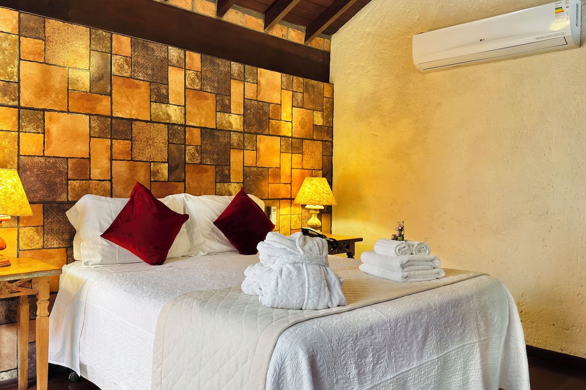 Quarto de hotel com parede de padrão em pedra, cama com roupa de cama branca e almofadas vermelhas, duas luminárias de cabeceira e uma pilha de toalhas sobre a cama.