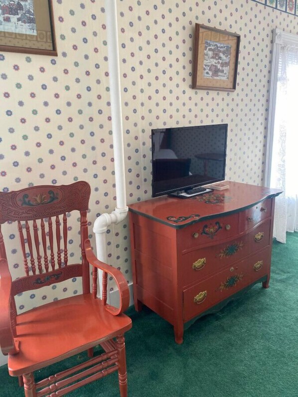 A room with a wooden chair, a red dresser with floral designs, a television, and patterned wallpaper.