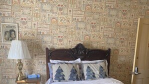 A bedroom with a patterned wallpaper, a wooden bed with a decorative headboard, a bedside table with a lamp, and a framed picture on the wall.