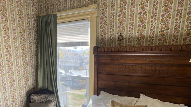 A bedroom with a floral-patterned bedspread, a wooden headboard, and a window with a view of the outdoors.